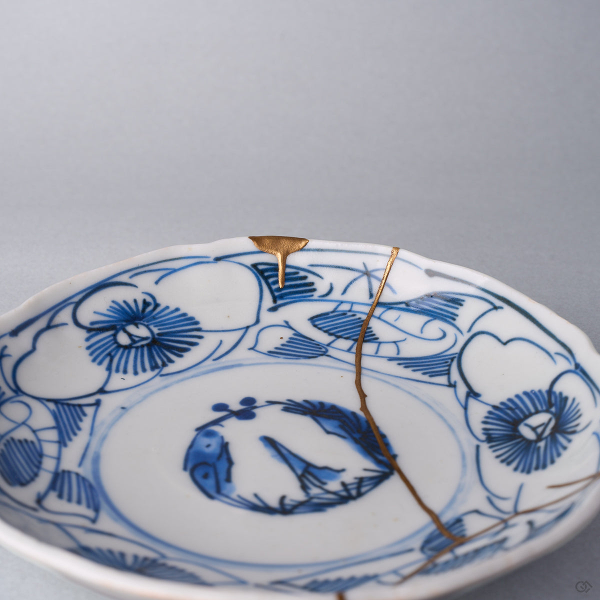 Kintsugi plate close-up in Ko-Imari porcelain, the natural urushi and 24K gold seam crossing the pine-and-plum roundel beside a gold-finished rim chip.