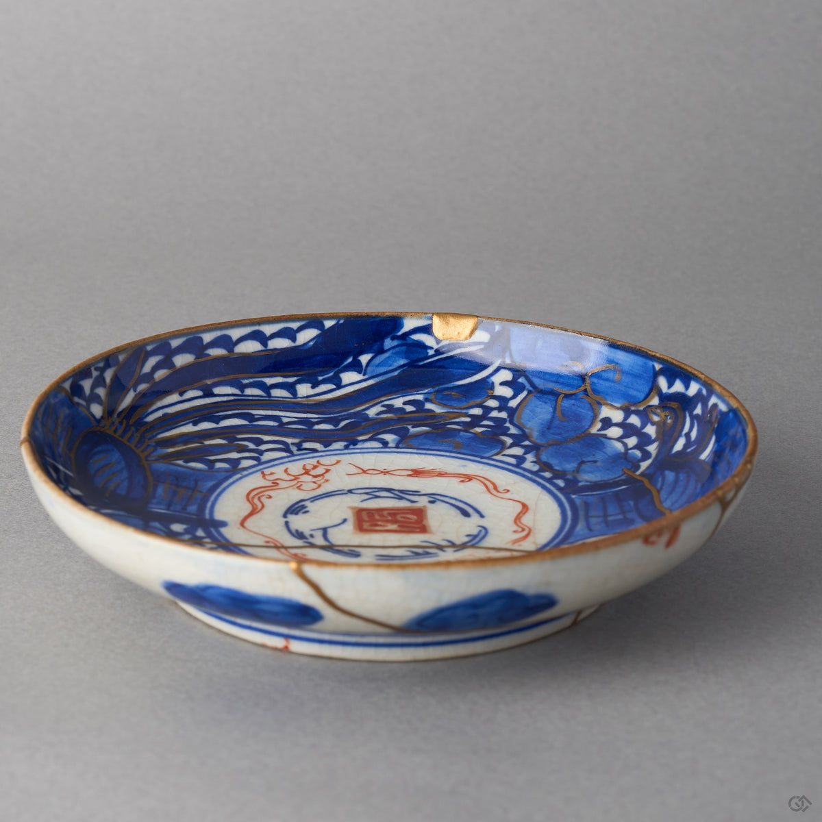 Kintsugi plate in Ko-Imari porcelain seen low across the well, rejoined with natural urushi, a 24K gold chip at the rim and a seam by the red seal, interior.