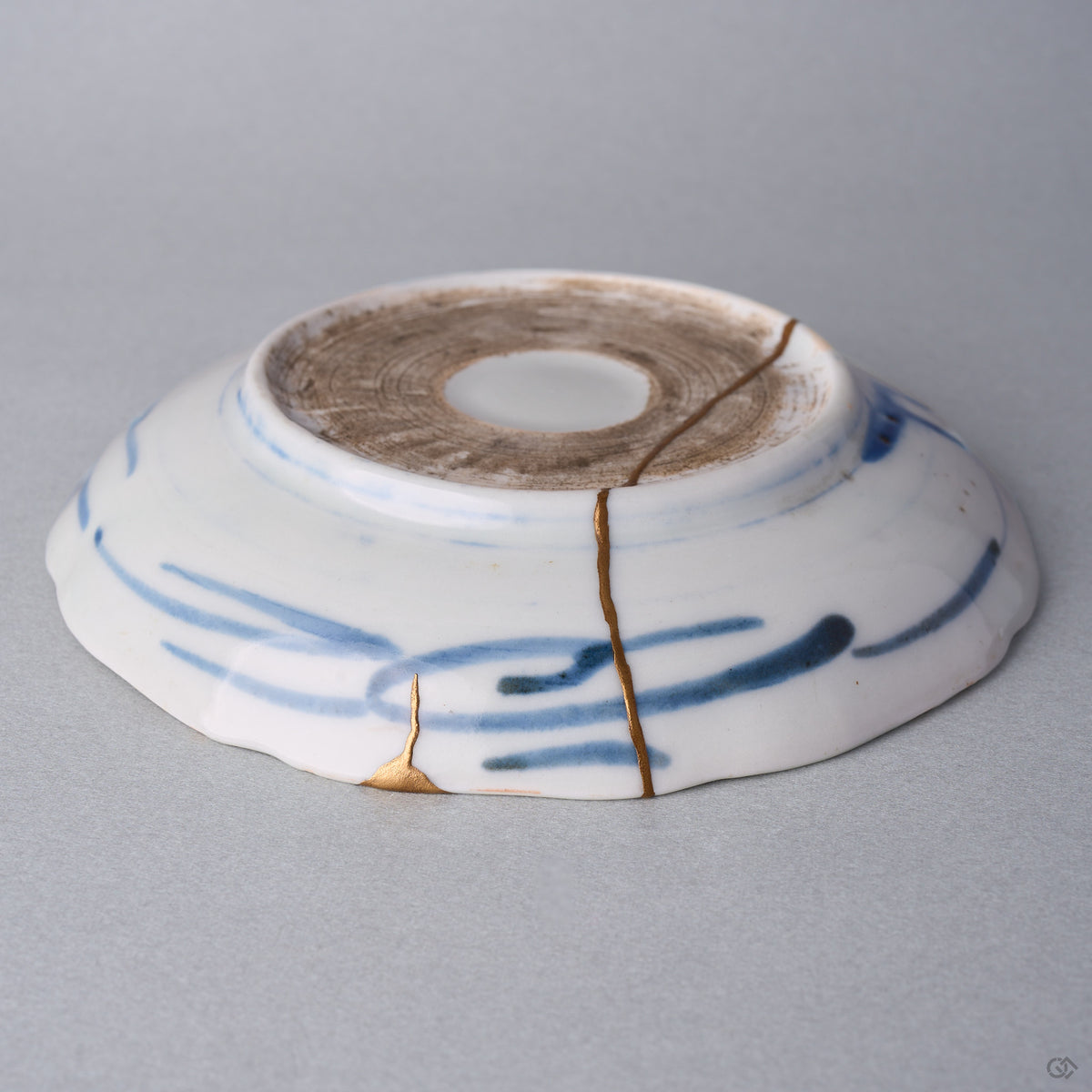 Kintsugi plate seen from below at an angle, natural urushi and 24K gold seams rising from a rim chip across the blue brushstrokes into the foot.