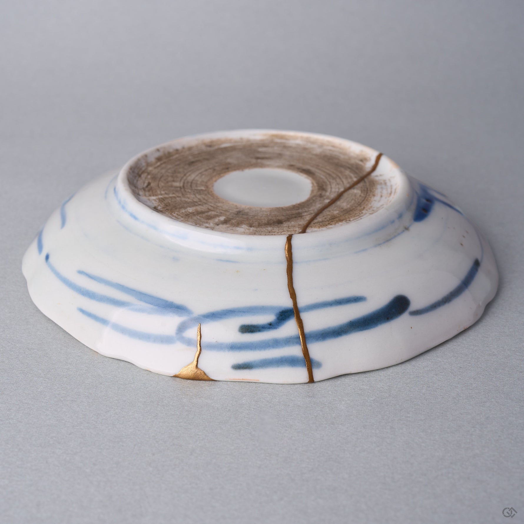 Kintsugi plate seen from below at an angle, natural urushi and 24K gold seams rising from a rim chip across the blue brushstrokes into the foot.