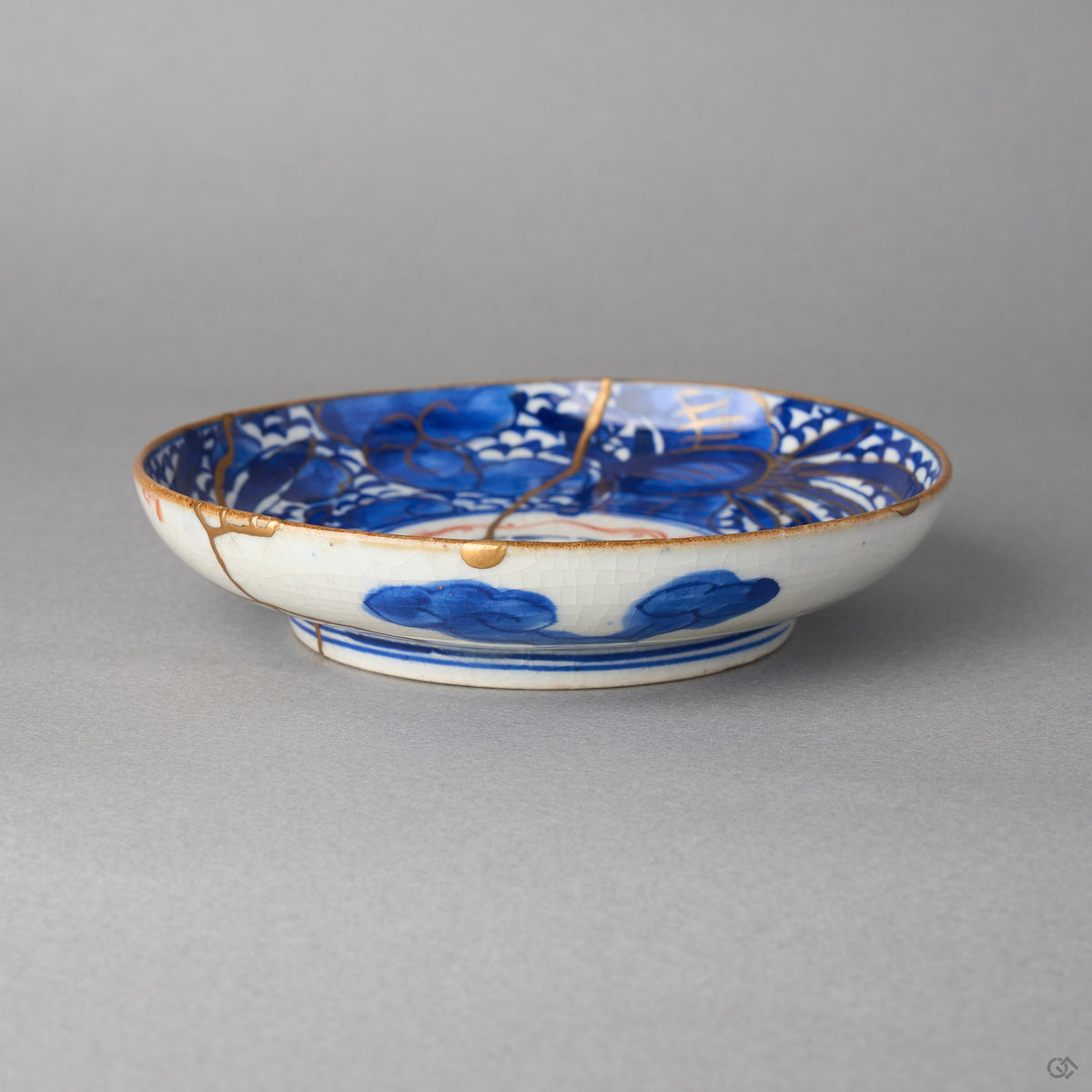 Kintsugi plate in Ko-Imari porcelain from the side, rejoined with natural urushi, a 24K gold seam dropping over the rim past a chip to the foot, side view.