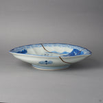 Kintsugi plate in Ko-Imari porcelain seen from the side on its low foot, rejoined with natural urushi, one 24K gold seam running from the rim down the far wall.
