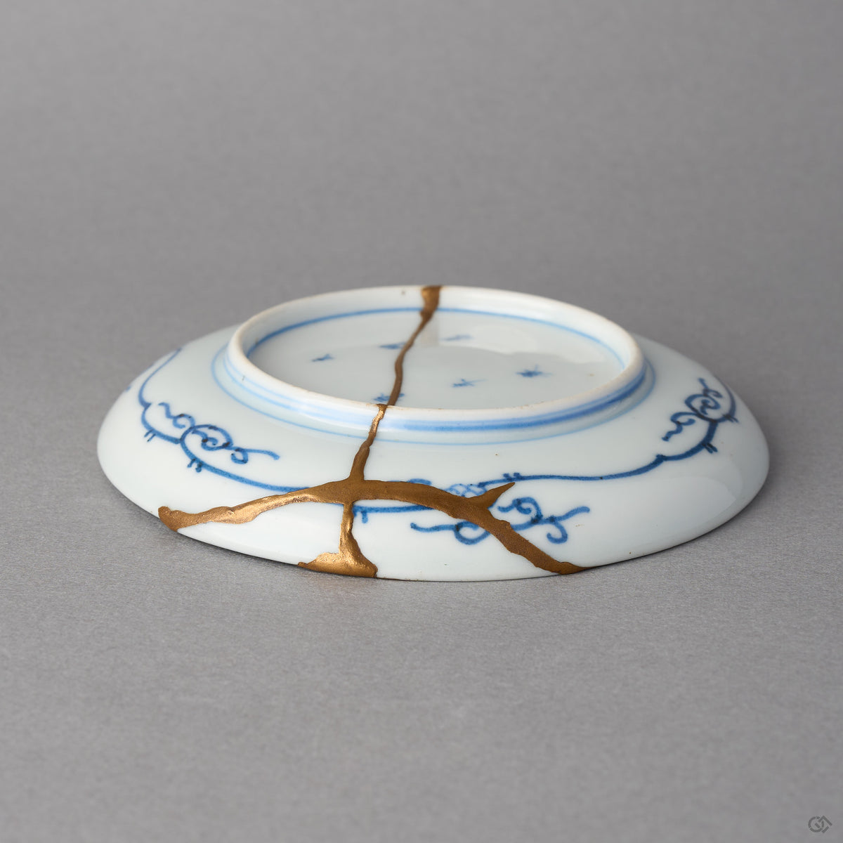Kintsugi plate in Ko-Imari porcelain seen from the back, rejoined with natural urushi, one 24K gold seam crossing the foot ring and branching toward the rim.