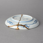 Kintsugi plate in Ko-Imari porcelain seen from the back, rejoined with natural urushi, one 24K gold seam crossing the foot ring and branching toward the rim.