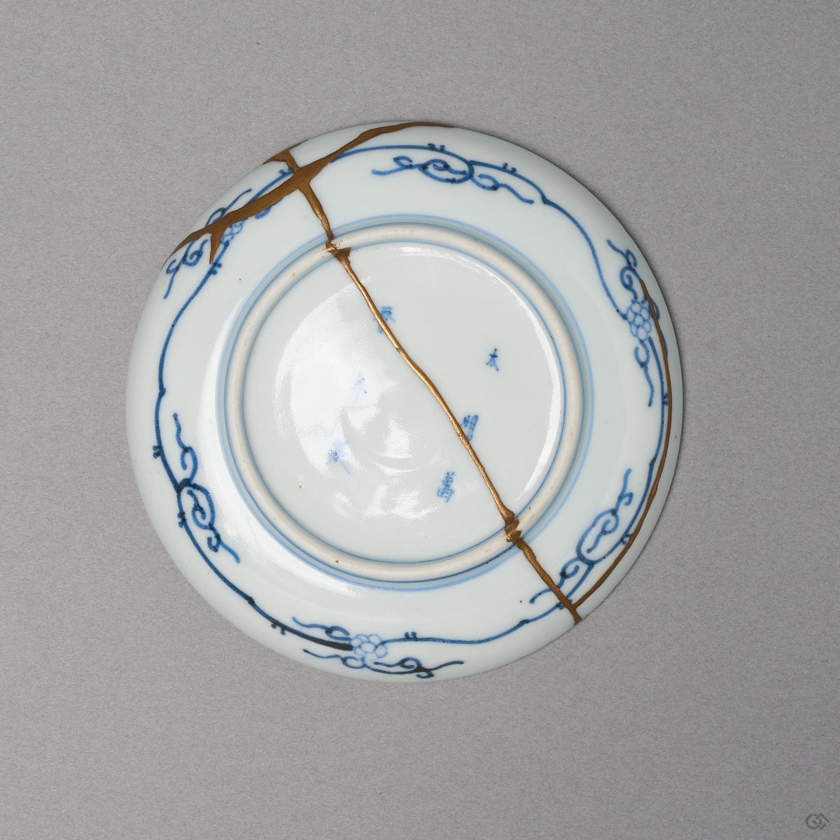 Kintsugi plate in Ko-Imari porcelain seen from below, rejoined with natural urushi, one 24K gold seam crossing the foot ring edge to edge between blue scrolls.