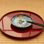 Kintsugi plate in Ko-Imari porcelain on a red lacquer tray with chopsticks, rejoined with natural urushi, its 24K gold seam and painted night sky seen in use.