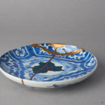 Kintsugi plate in Ko-Imari porcelain at a low angle, rejoined with natural urushi, the 24K gold seam running from the rim through the urushi-e sky and stars.