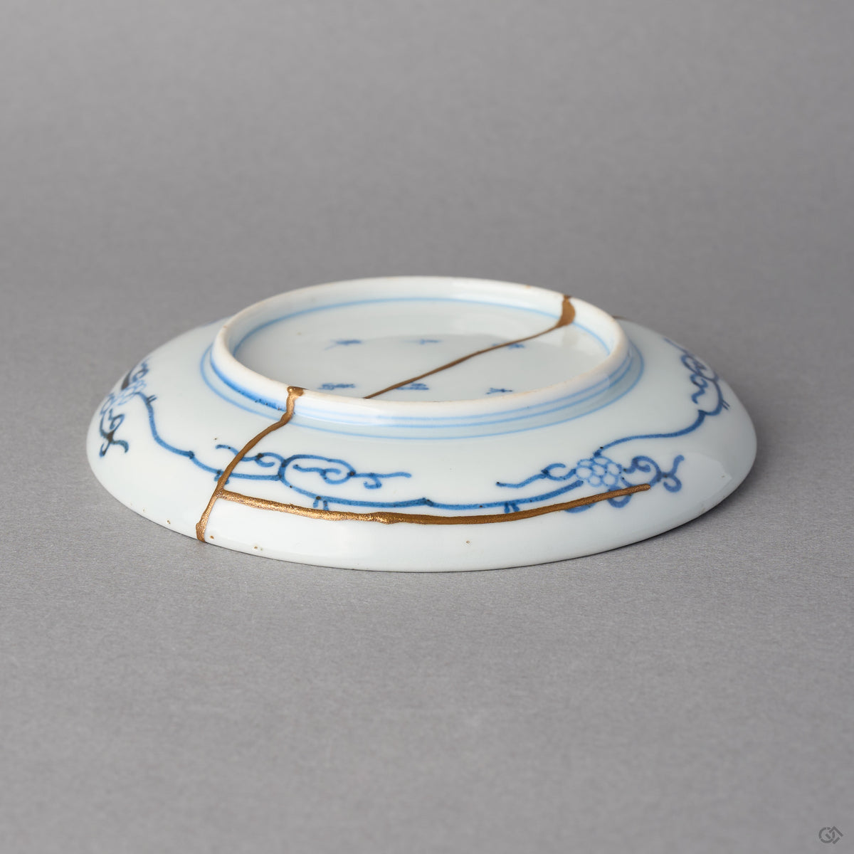 Kintsugi plate in Ko-Imari porcelain tilted to its underside, rejoined with natural urushi, a 24K gold seam along the blue karakusa border and across the foot.