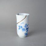 Kintsugi sake cup in thin Arita porcelain, rejoined with natural urushi, one 24K gold seam sweeping from the rim above the blue vine, front view.