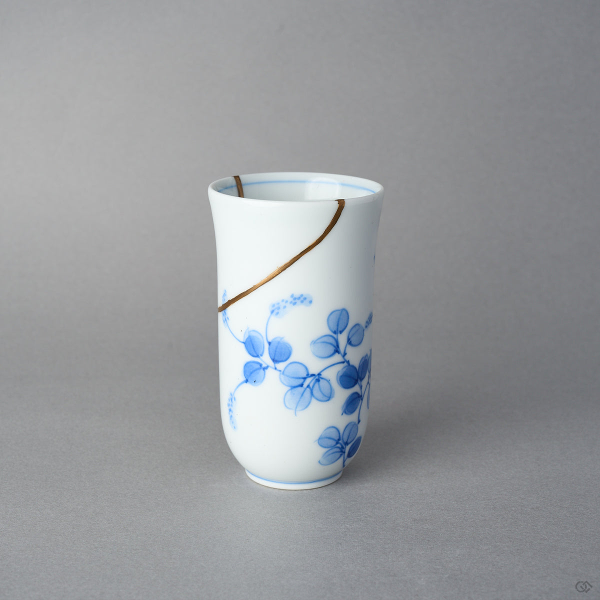 Kintsugi sake cup in thin Arita porcelain, rejoined with natural urushi, one 24K gold seam sweeping from the rim above the blue vine, front view.
