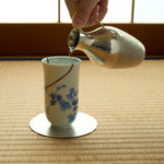 Kintsugi sake cup in Arita porcelain on tatami, sake poured from a hammered tin flask, rejoined with natural urushi, one 24K gold arc above the blue vine.