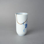 Kintsugi sake cup in Arita porcelain, rejoined with natural urushi, the 24K gold seam dipping from the rim in one arc above the blue leaves, side view.