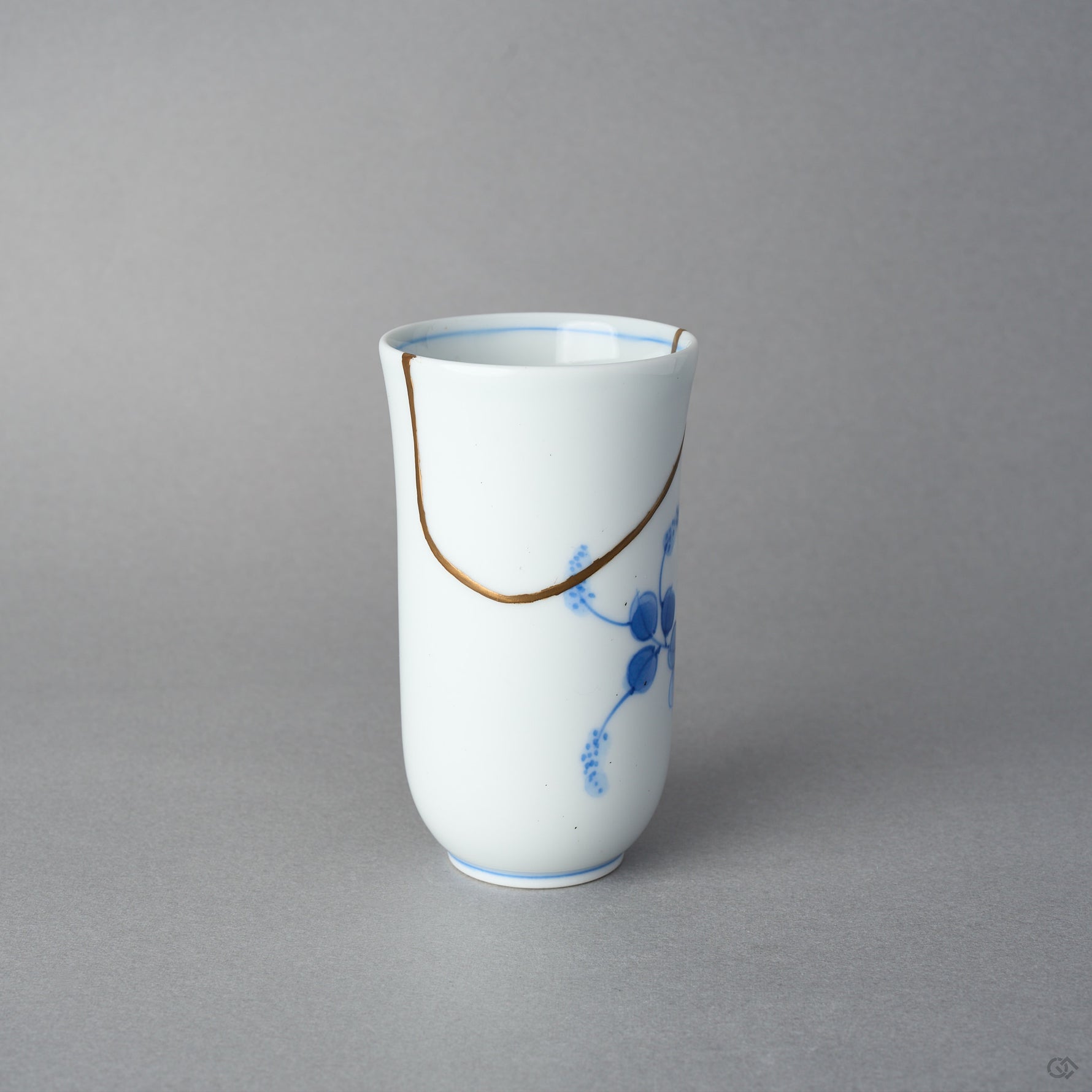 Kintsugi sake cup in Arita porcelain, rejoined with natural urushi, the 24K gold seam dipping from the rim in one arc above the blue leaves, side view.