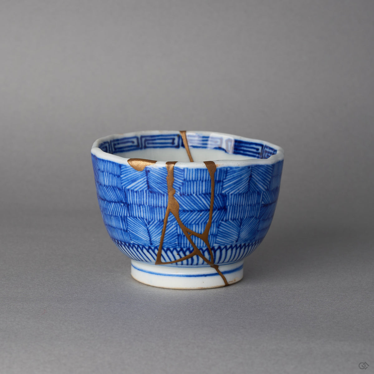 Kintsugi sake cup in checkered blue Ko-Imari porcelain, rejoined with natural urushi, one 24K gold seam descending from a filled rim chip, front view.