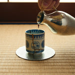 Kintsugi sake cup in Ko-Imari porcelain receiving sake from a tin flask on tatami, its natural urushi and 24K gold seam facing the viewer.
