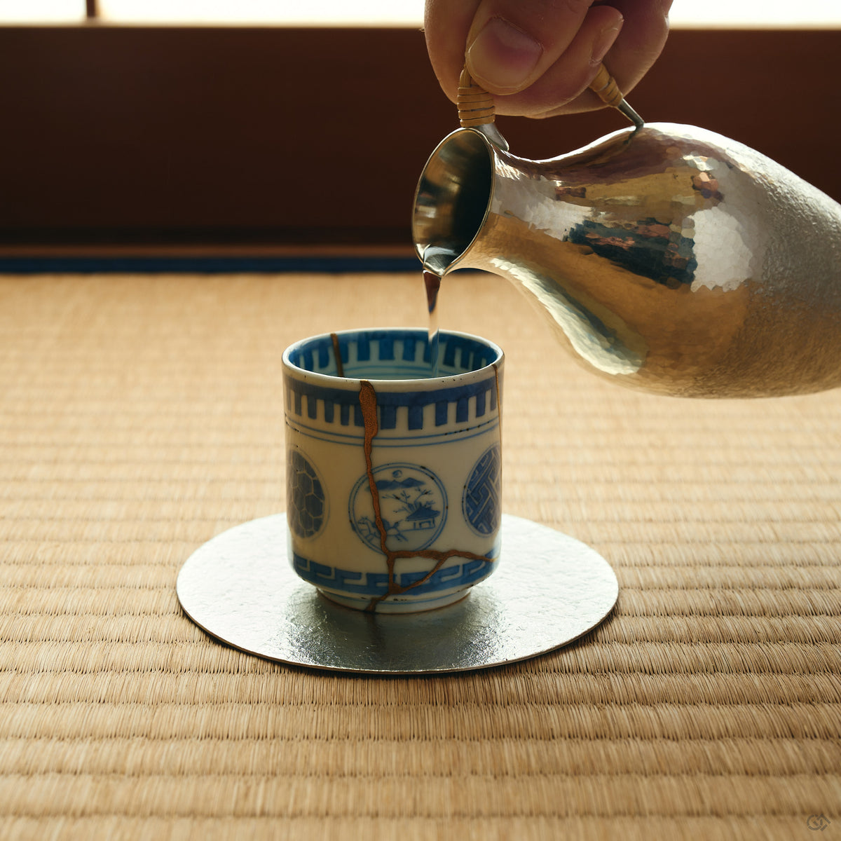 Kintsugi sake cup in Ko-Imari porcelain receiving sake from a tin flask on tatami, its natural urushi and 24K gold seam facing the viewer.
