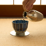 Kintsugi sake cup in Ko-Imari porcelain filled from a hammered tin flask on a tatami mat, rejoined with natural urushi, 24K gold seams on the front wall.