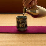 Kintsugi sake cup in Ko-Imari porcelain on tatami, sake poured from a hammered tin flask, the rim rebuilt in natural urushi under a 24K gold sparrow.