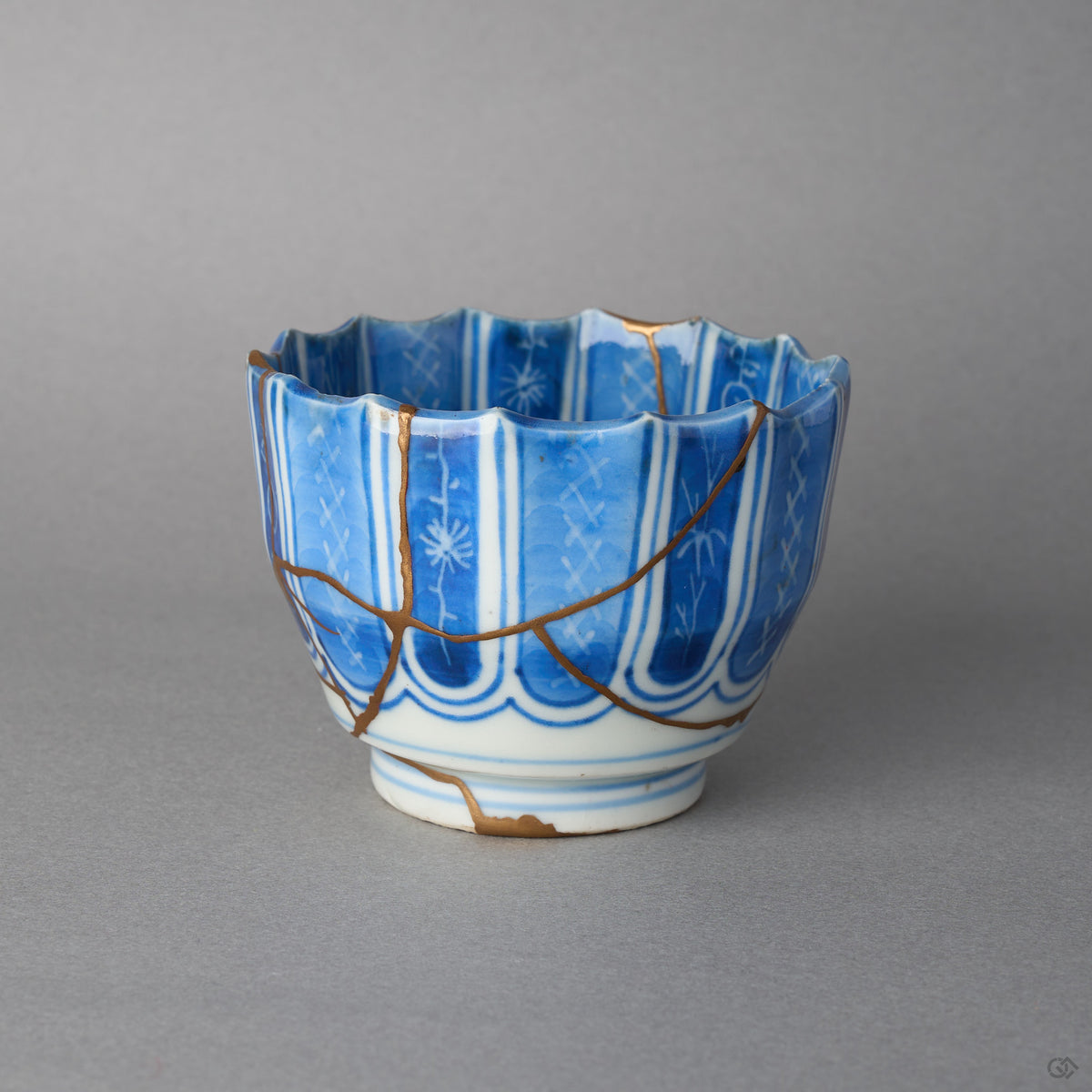 Kintsugi sake cup in Ko-Imari porcelain with a scalloped rim, rejoined with natural urushi, one 24K gold seam running from rim to foot, side view.
