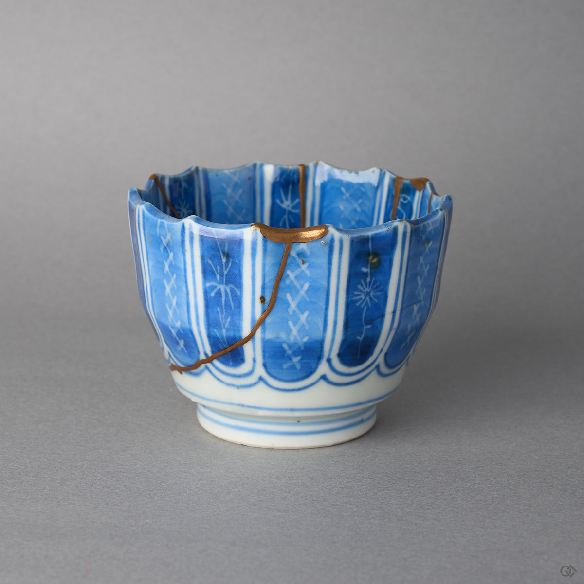 Kintsugi sake cup in Ko-Imari porcelain, rejoined with natural urushi, a 24K gold seam branching from a filled rim chip across the blue columns, side view.