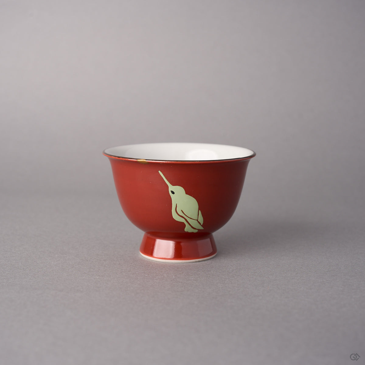 Kintsugi sake cup in red Kyoto ware porcelain, rejoined with natural urushi, one 24K gold point at the rim above a pale hummingbird looking up, front view.