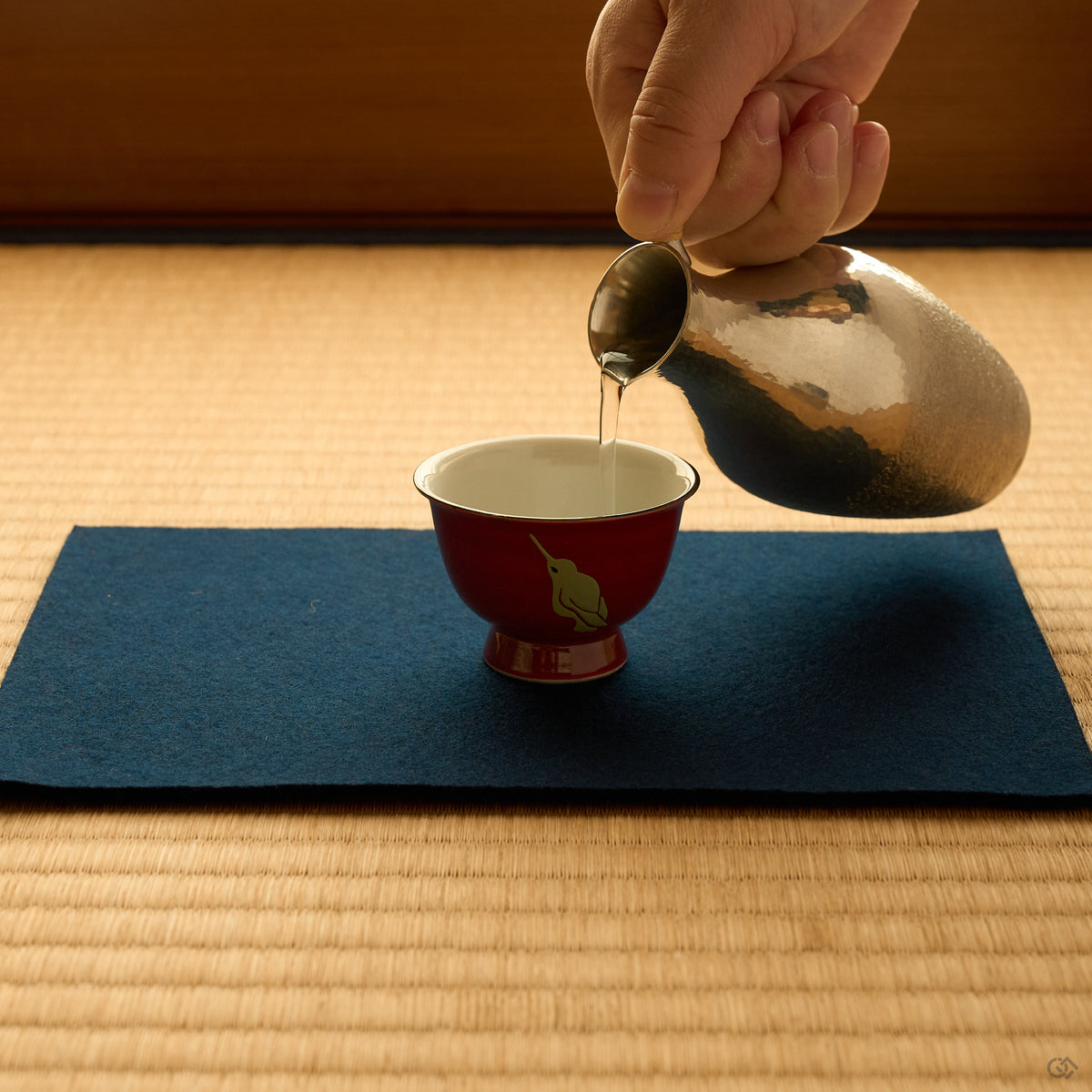 Kintsugi sake cup in red Kyoto ware porcelain on indigo felt and tatami, sake poured from a hammered tin flask, a hummingbird painted in urushi, in-use view.