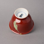 Kintsugi sake cup in red Kyoto ware porcelain tipped on its rim, restored with natural urushi, the 24K gold point beside the perched hummingbird, side view.