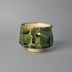Kintsugi sake cup in faceted Oribe green pottery, a natural urushi and 24K gold seam forking below the rim and descending the facet to the foot, front view.