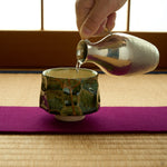 Kintsugi sake cup in Oribe green pottery on a magenta runner over tatami as sake is poured from a hammered tin flask, urushi and 24K gold on its facets.