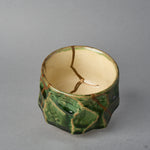 Kintsugi sake cup in Oribe green pottery viewed from above at an angle, natural urushi and 24K gold seams entering the ivory well from three sides.
