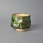 Kintsugi sake cup in Oribe ware seen from the side, a natural urushi and 24K gold seam slanting across a milky pool of glaze on the copper-green facets.