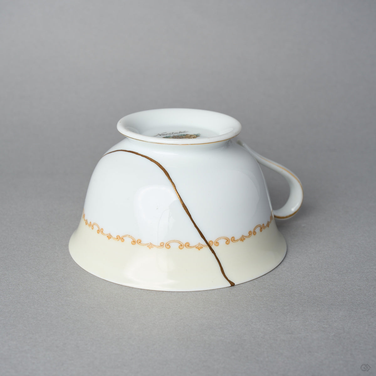 Kintsugi tea cup in Early Noritake porcelain, inverted side view: the 24K gold urushi seam runs from the foot across the ivory band, handle at right.