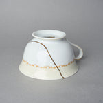 Kintsugi tea cup in Early Noritake porcelain, inverted side view: the 24K gold urushi seam runs from the foot across the ivory band, handle at right.