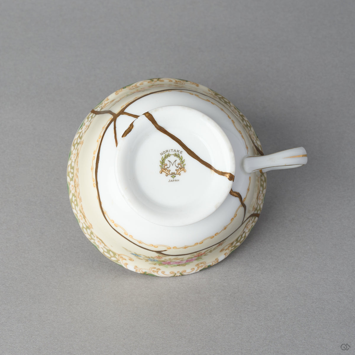Kintsugi tea cup in Early Noritake porcelain seen from below, natural urushi and 24K gold seams crossing the base and foot ring, bottom view.