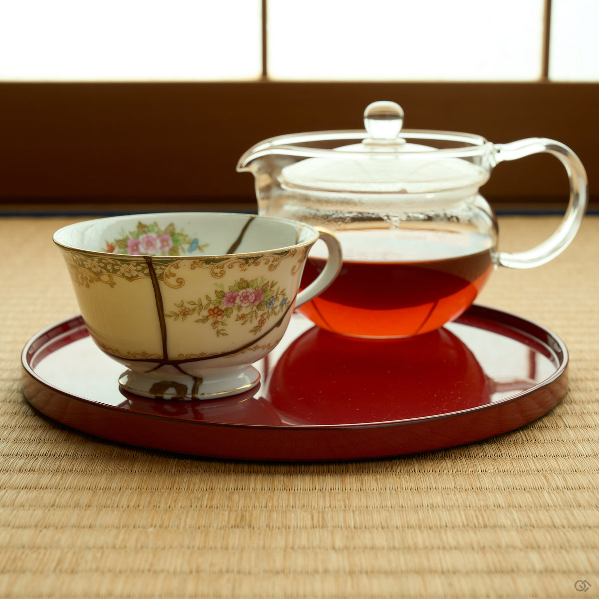 Kintsugi tea cup in Early Noritake porcelain on a red lacquer tray beside a glass teapot on tatami, natural urushi and 24K gold seams meeting at its foot.