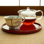 Kintsugi tea cup in Early Noritake porcelain on a red lacquer tray beside a glass teapot on tatami, natural urushi and 24K gold seams meeting at its foot.