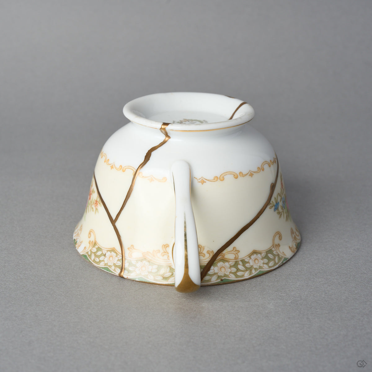 Kintsugi tea cup in Early Noritake porcelain resting inverted, a natural urushi and 24K gold seam forking across the gilt scroll band, side view.