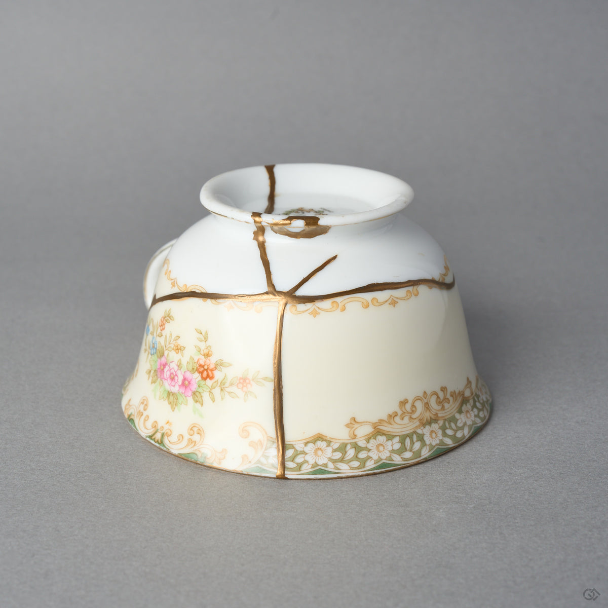Kintsugi tea cup in cream Early Noritake porcelain resting inverted, natural urushi and 24K gold seams crossing the gilt scroll band to the rim, side view.