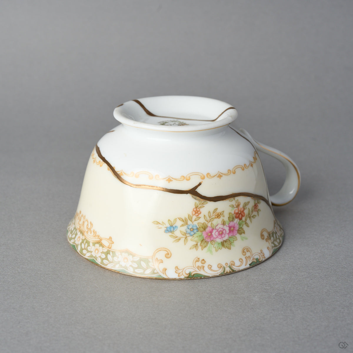 Kintsugi tea cup in Early Noritake porcelain resting inverted, one natural urushi and 24K gold line curving along the scroll band above the roses, side view.
