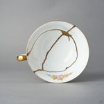 Kintsugi tea cup in Early Noritake porcelain seen from directly above, natural urushi and 24K gold seams gathering into a star in the white well, top view.