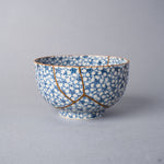 Kintsugi tea cup in Kyoto ware: a branching seam of natural urushi and 24K gold descends the packed cherry-blossom porcelain, front view.