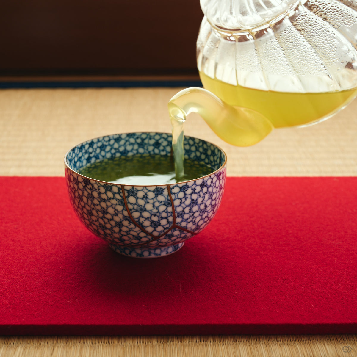 Kintsugi tea cup in Kyoto ware receiving green tea on a red felt mat, its urushi and 24K gold seam crossing the blossom pattern.