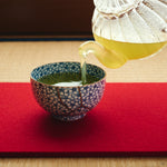Kintsugi tea cup in Kyoto ware receiving green tea on a red felt mat, its urushi and 24K gold seam crossing the blossom pattern.