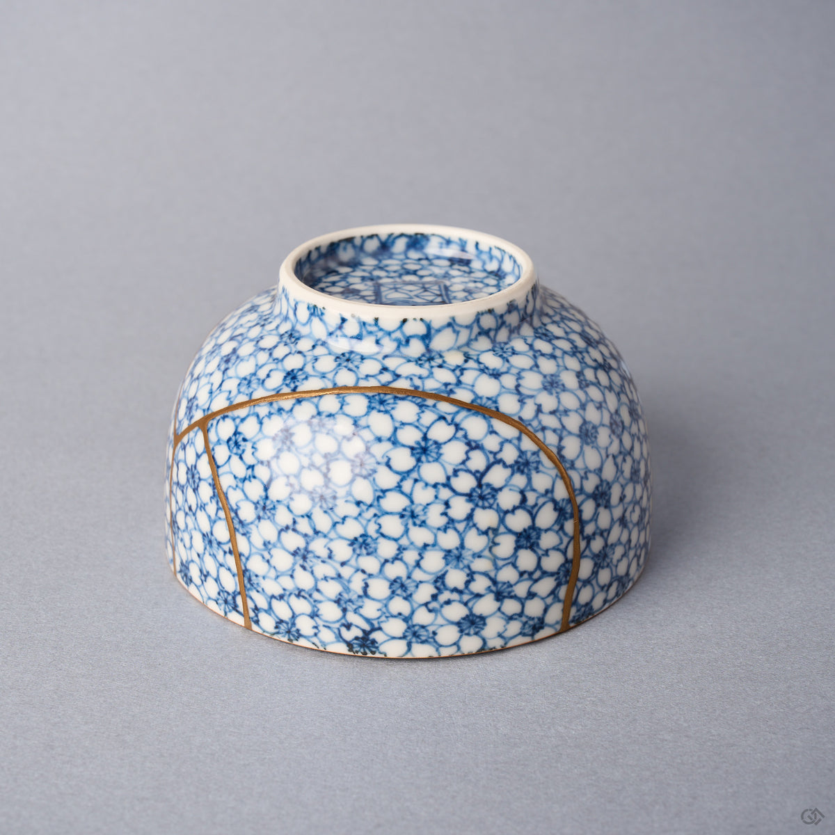 Kintsugi tea cup inverted: a long urushi and 24K gold line arcs around the lower body across the cherry-blossom porcelain.