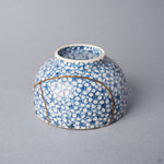 Kintsugi tea cup inverted: a long urushi and 24K gold line arcs around the lower body across the cherry-blossom porcelain.