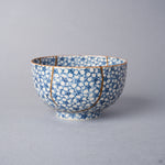 Kintsugi tea cup side view: two urushi and 24K gold lines descend the cherry-blossom wall from the iron-brown rim.