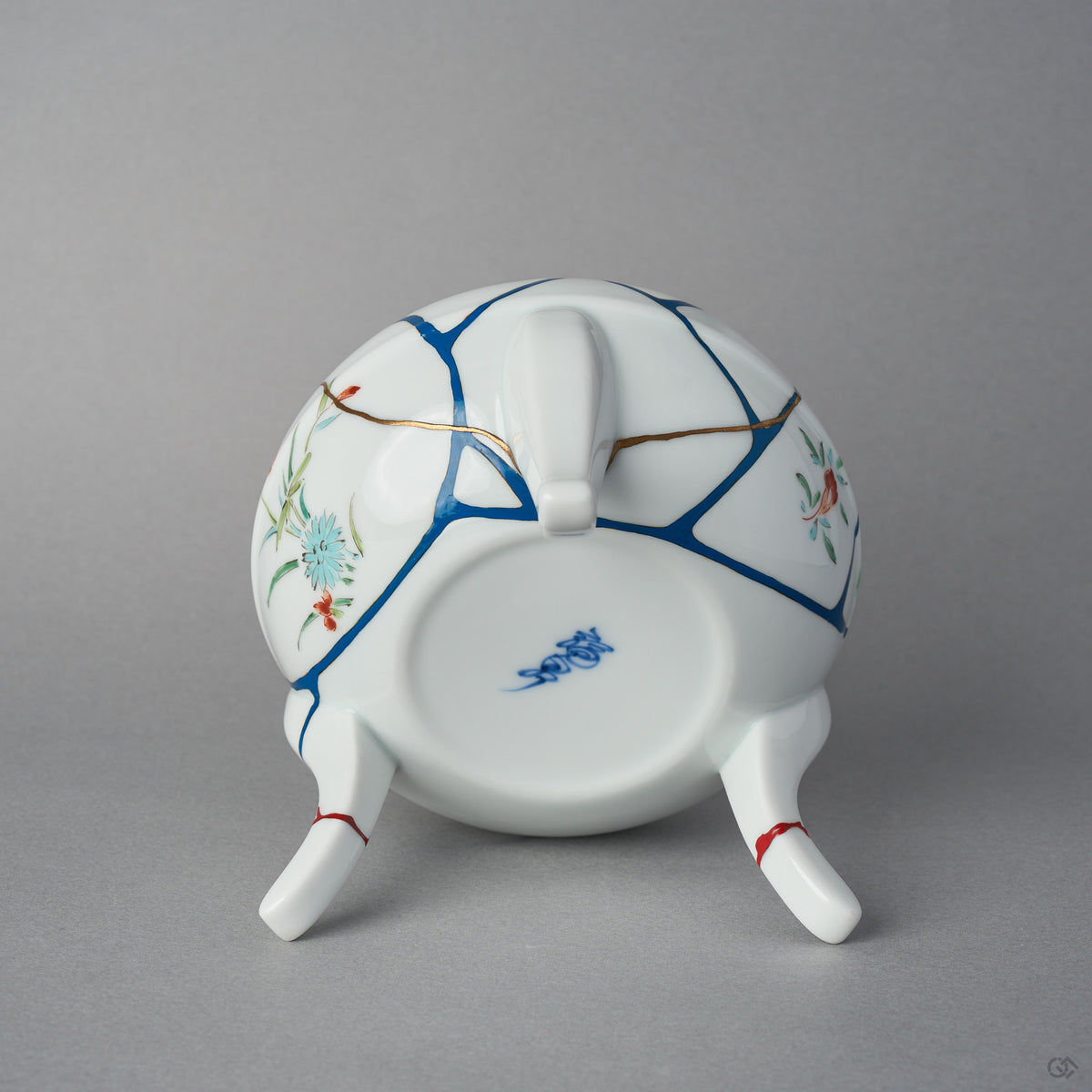 Underside of the Arita lidded bowl body showing the blue potter's mark, blue urushi lines with 24K gold edges, and the three restored feet ringed in vermilion colored urushi.