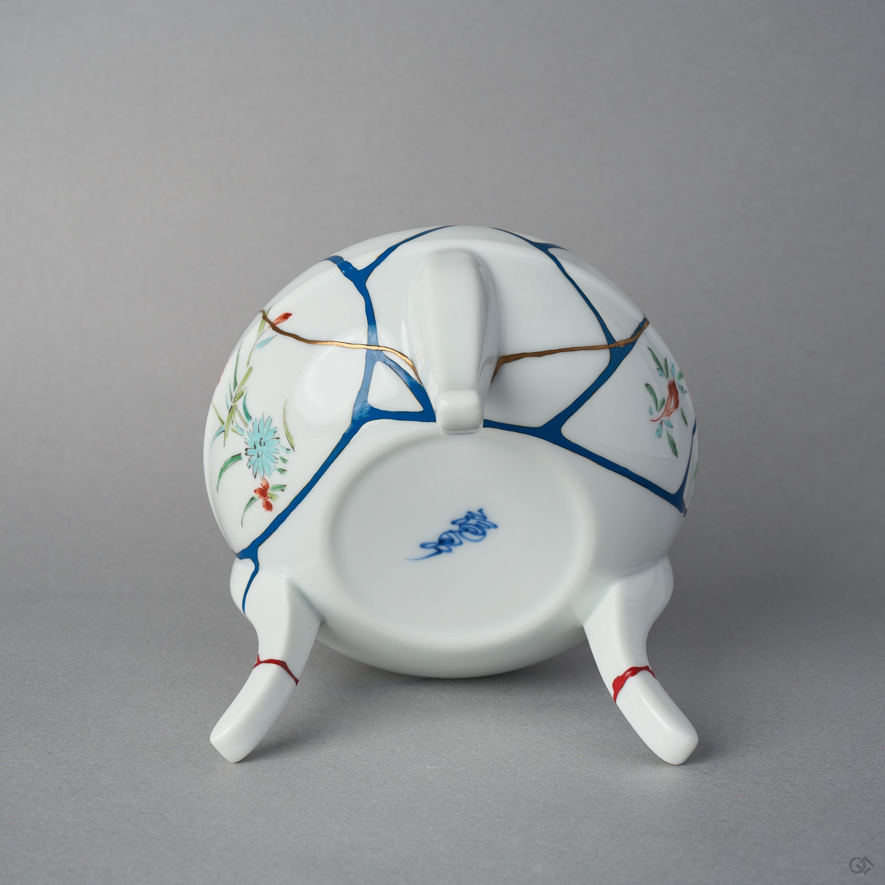 Underside of the Arita lidded bowl body showing the blue potter's mark, blue urushi lines with 24K gold edges, and the three restored feet ringed in vermilion colored urushi.