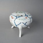 Front view of an Arita ware lidded bowl on three feet, white porcelain with painted flowers and butterflies, blue urushi lines, 24K gold kintsugi seams, and swan maki-e on the lid.