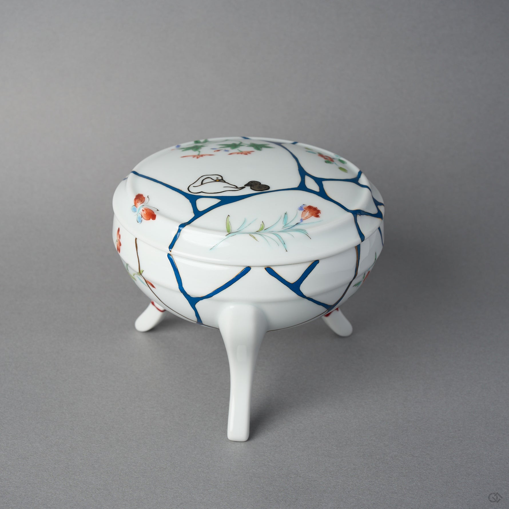 Front view of an Arita ware lidded bowl on three feet, white porcelain with painted flowers and butterflies, blue urushi lines, 24K gold kintsugi seams, and swan maki-e on the lid.